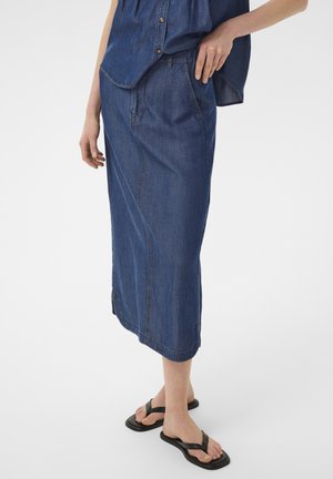 Žena nosi modrú denimovú midi sukňu a zodpovedajúci bezrukávový top na gombíky, stojí v čiernych žabkách na jednoduchom pozadí.