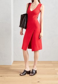 Robe rouge côtelée avec un décolleté en V et une fente latérale, associée à un sac à main noir tressé et des mules à talons blocs ouverts. Texture lisse, coupe ajustée.