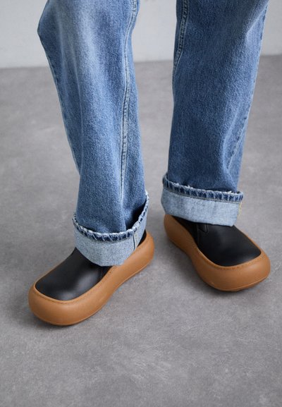 Melni slip-on apavi ar noapaļotu brūnu gumijas zoli, valkāti ar uzlocītām zilām džinsu biksēm. Apaviem ir mīksta tekstūra un gluda apdare.