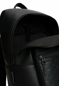 Sac à dos noir en cuir texturé et tissu lisse, comprenant un compartiment zippé et des détails en relief sur la poche avant.