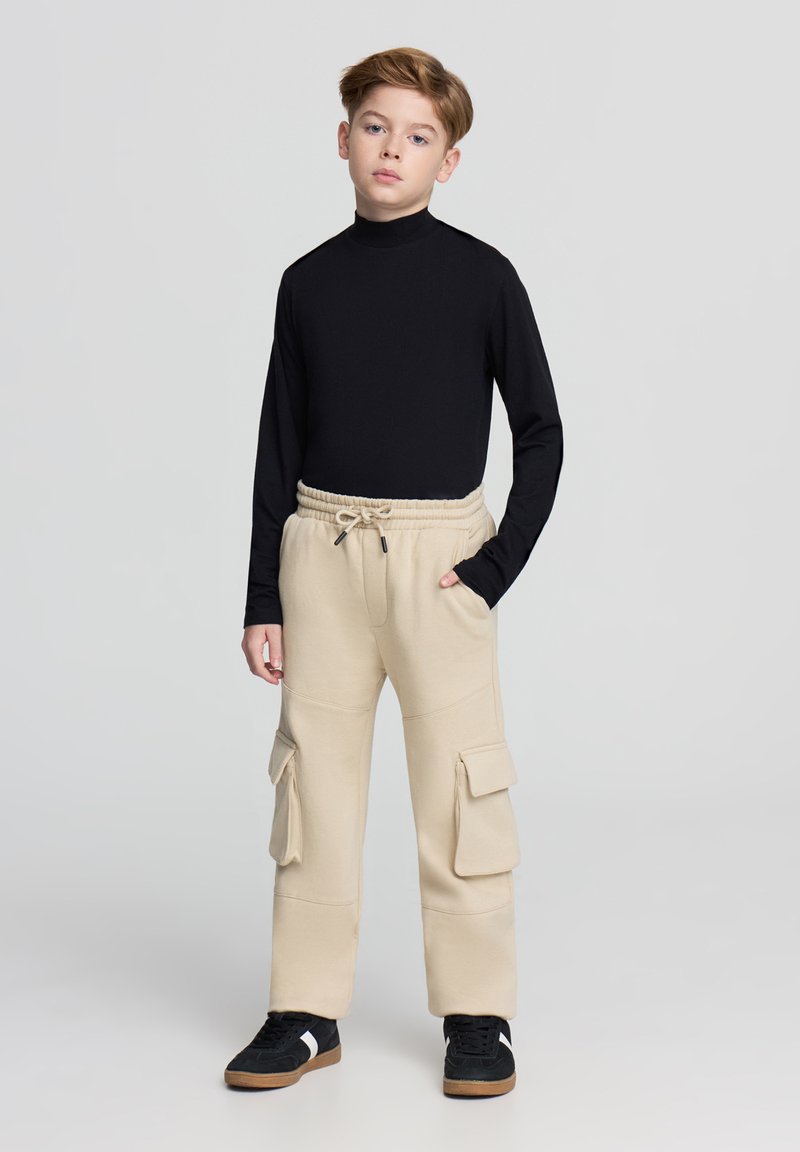Maglione nero a collo alto con maniche lunghe, pantaloni cargo beige in felpa con vita elastica e grandi tasche laterali, abbinati a sneakers nere con dettagli bianchi.