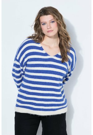 Kvinde med bølget blondt hår iført blå- og hvidstribet sweater og sorte bukser, stående mod en hvid baggrund med hænderne bag ryggen.