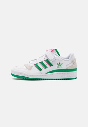 Zapatilla blanca Adidas con rayas verdes, suela verde, detalles de ante beige y una tira de velcro sobre un fondo claro y liso.