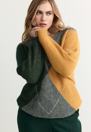 Blonde Frau trägt einen color-block Pullover mit Senf-, Dunkelgrün- und Grautönen und posiert mit den Händen nahe ihrem Gesicht.