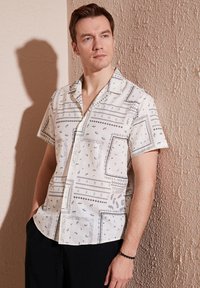 Camisa de manga corta y botones en crema claro con patrones geométricos negros, que presenta un corte relajado y un diseño de cuello sencillo.