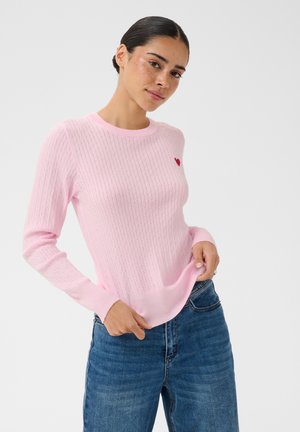 Femme portant un pull en maille torsadée rose clair avec un petit cœur rouge sur la poitrine et un jean bleu, posant avec une main dans la poche.