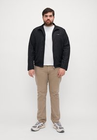 Giacca nera leggera con zip frontale, indossata su una camicia bianca, abbinata a pantaloni beige e sneakers. Il design è semplice e funzionale.