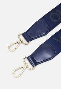 Tmavě modrý látkový popruh tašky s koženými akcenty a zlatými kovovými otočnými sponami na obou koncích, s částečně viditelným textem v embosovaném písmu.