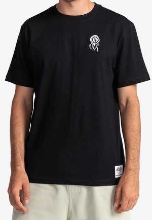 Homme portant un t-shirt noir avec un petit graphisme d'un arbre blanc avec des racines en haut à gauche de la poitrine et une étiquette logo rectangulaire près de l'ourlet.