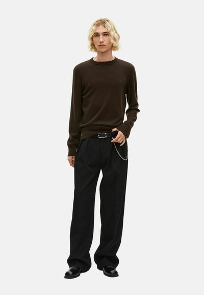 Joven con cabello rubio hasta los hombros, vistiendo un suéter marrón oscuro, pantalones anchos negros con cinturón y cadena, y zapatos negros.
