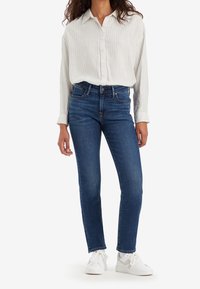 Femme portant une chemise blanche à fines rayures boutonnée, rentrée dans un jean bleu taille haute et des baskets blanches, debout sur un fond blanc.