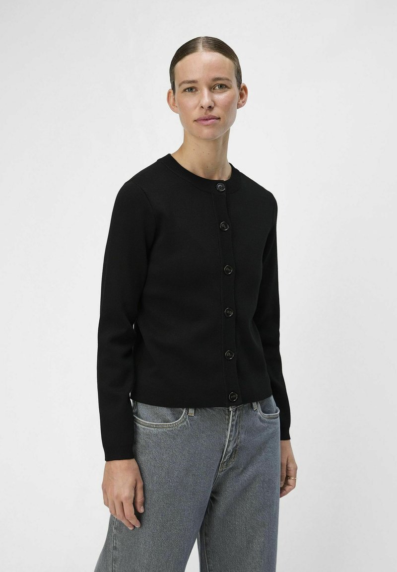 Cardigan noir avec un col rond, présentant une rangée de six boutons sur le devant. Coupe classique avec des manches longues, associé à un jean gris clair.