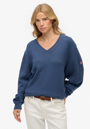 V-hals sweater i dyb blå strikstof med ribbede ærmer og kant, med et lille logopatchet på ærmet. Parret med lyse bukser.