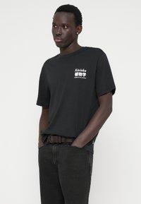 Μαύρο βαμβακερό μπλουζάκι με λευκό εκτυπωμένο κείμενο και γραφικά, συνδυασμένο με σκούρα τζιν. Απλή, χαλαρή εφαρμογή και στρογγυλή λαιμόκοψη.