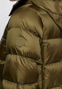 Olivově zelená péřová bunda s prošitým vzorem, hladkou texturou a kulatou logovou nášivkou na rukávu. Má vysoký límec a lesklý povrch.
