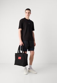 T-shirt in cotone nero con un piccolo logo, abbinato a pantaloni corti neri con accenti blu. Tiene una borsa in tela nera con un'etichetta rossa.