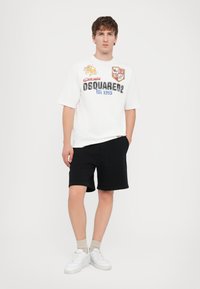 Camiseta blanca con estampados gráficos y texto, combinada con pantalones cortos negros. El modelo lleva zapatillas blancas y calcetines beige. Diseño simple y casual.