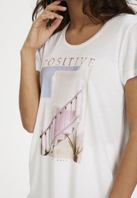 Mujer vestida con una camiseta blanca con una escalera en tonos pastel, una planta y la palabra "POSITIVO" encima de la imagen.