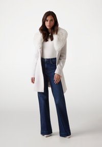 Manteau blanc avec un col en fausse fourrure, associé à un jean large taille haute bleu foncé. Tissu lisse, design structuré et boutons dorés.