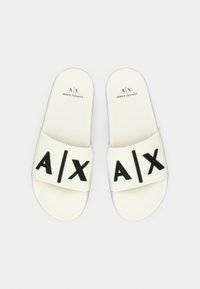 Chinelos brancos com uma tira texturada. Grandes letras pretas "A/X" impressas na tira. Design minimalista com sola plana.