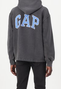 Felpa grigia con cappuccio e logo blu "GAP", morbida al tatto, vestibilità ampia, polsini e orlo a coste. Abbinata a pantaloni scuri.