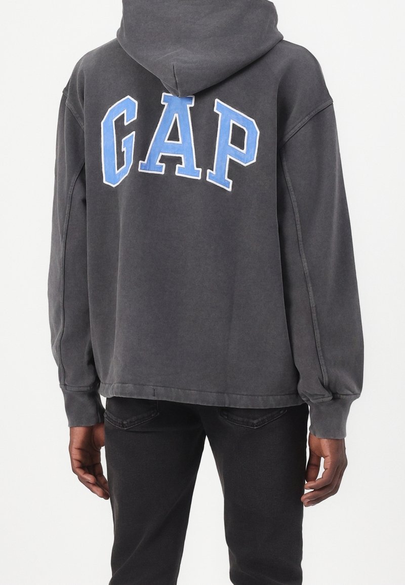 Felpa grigia con cappuccio e logo blu "GAP", morbida al tatto, vestibilità ampia, polsini e orlo a coste. Abbinata a pantaloni scuri.