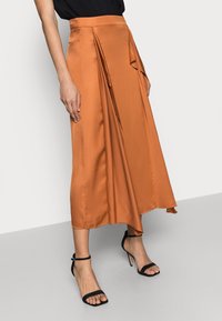 Malli joka käyttää eleganttia, hulmuavaa oranssia hametta, jossa on poimutuksia, yhdistettynä mustiin remmikenkiin korkealla korolla. Neutraali tausta korostaa asua.