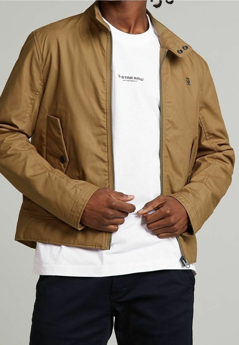 Brun, lätt jacka med dragkedja fram, sidofickor och ståkrage. Bärs över en vit t-shirt. Slät textur, avslappnad design.