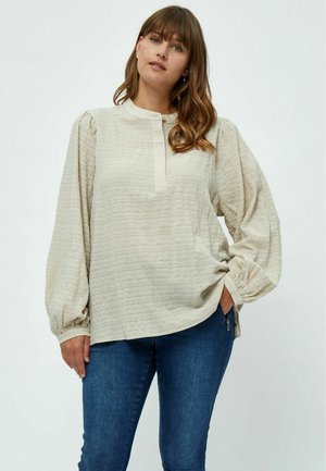 Cream-colored, textured blouse with puffed sleeves, button placket, and ribbed detailing, paired with dark blue fitted jeans.