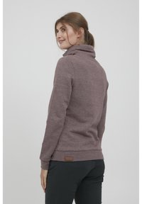 Lila tröja med hög krage, texturerat tyg och en logotypapplikation vid nederkanten. Har en avslappnad passform och långa ärmar.