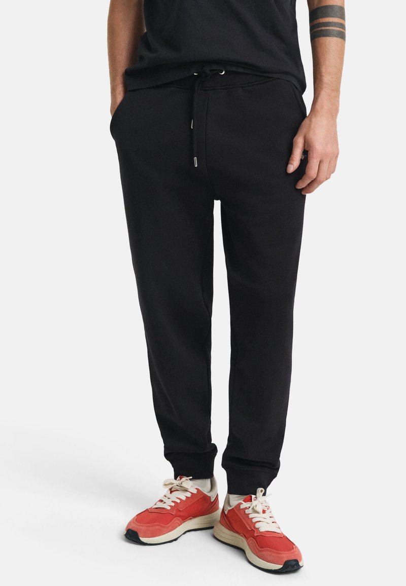 Sorte sweatpants med elastisk talje og snoretræk. Materialet ser blødt ud. Modellen har røde sneakers på med hvide accenter og beige såler.