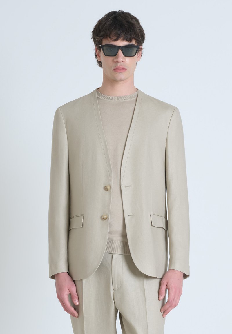 Blazer beige con due bottoni, rever e tasche laterali. Indossato sopra un maglione beige abbinato. Il modello indossa occhiali da sole scuri.