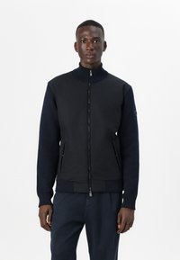 Μαύρο bomber μπουφάν με ψηλό γιακά, φερμουάρ μπροστά και ριμπ μανσέτες. Το πάνω μέρος διαθέτει λείο ύφασμα, ενώ το κάτω μέρος περιλαμβάνει υφή και λεπτομέρειες.