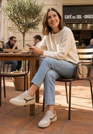 Mujer joven con suéter blanco y jeans sentada en una mesa de café al aire libre, sosteniendo un teléfono, sonriendo, con una taza de café cerca y dos hombres al fondo.