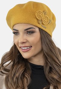 Geel wollen baret met drie opgerolde bloemenaccenten. Gladde textuur met een iets slungelige vorm, die een zwart outfit aanvult.
