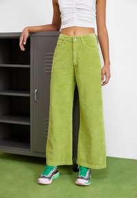 Pantalones de pana verde de pierna ancha con una superficie texturizada, que cuentan con un cierre de botón y bolsillos frontales, combinados con zapatillas de colores múltiples.