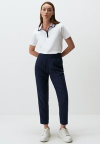 Witte polo met een zwarte ritssluiting bij de kraag, gecombineerd met marineblauwe tapered broek en witte sneakers. De outfit heeft een minimalistisch ontwerp en strakke lijnen.