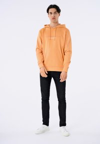 Orange hoodie med framficka och dragsko, prydd med vit text "Copenhagen Lindbergh." Kombinerad med svarta slim-fit jeans och vita sneakers.