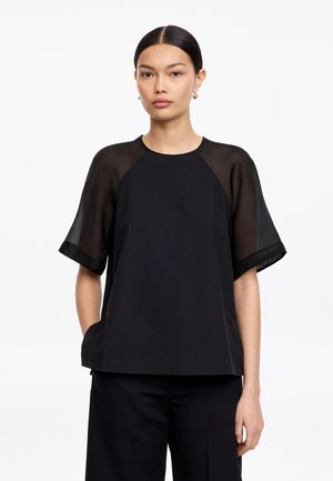 Femme aux cheveux raides et foncés portant un haut noir à manches courtes transparentes et un pantalon noir à jambes larges sur fond blanc.
