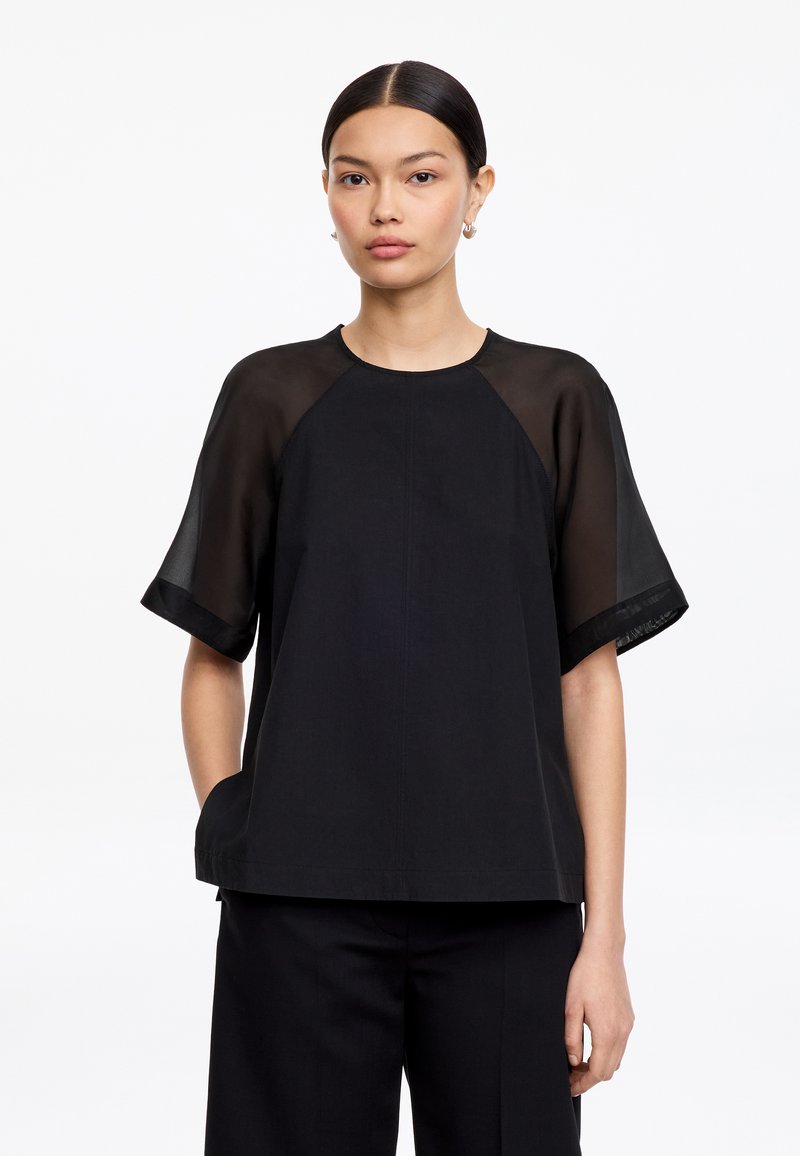 Femme aux cheveux raides et foncés portant un haut noir à manches courtes transparentes et un pantalon noir à jambes larges sur fond blanc.