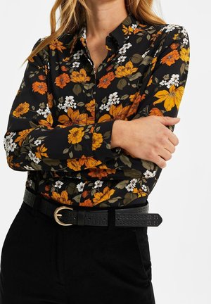 Donna che indossa una camicia nera a fiori con bottoni, con fiori arancioni, gialli e bianchi, abbinata a pantaloni neri e una cintura nera con fibbia dorata.