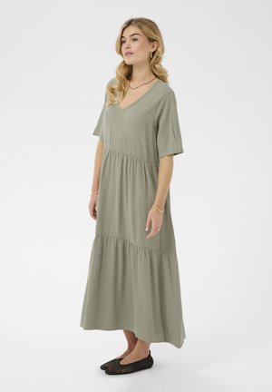 Donna con capelli biondi ondulati che indossa un abito midi verde salvia scollato a V e scarpe basse nere intrecciate, in piedi davanti a uno sfondo bianco semplice.