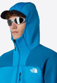 Modrá kapuce s zipem, texturovaná tkanina a logo The North Face na hrudi. Doplněno bílými slunečními brýlemi a černou čepicí.
