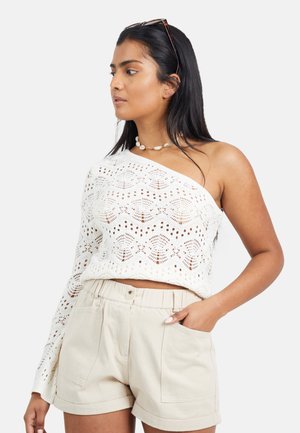 Kobieta w białym, jednostronnym swetrze na jedno ramię wykonanym na szydełku oraz beżowych, wysokiego stan szortach, z ręką w kieszeni i naszyjnikiem z muszli, patrząca na bok.