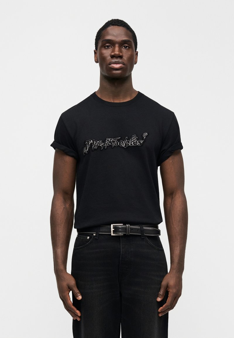 T-shirt en coton noir avec un graphique texturé et stylisé blanc sur le devant. Associé à un denim foncé et une ceinture noire.