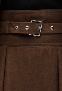 Καφέ υφασμάτινη ζώνη με ασημένια ορθογώνια πόρπη στερεωμένη γύρω από τη μέση του καφέ ρούχου που ταιριάζει.