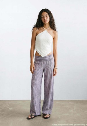 Giovane donna con lunghi capelli ricci che indossa un top bianco all’halter, pantaloni larghi a fantasia, sandali neri, in piedi davanti a uno sfondo uniforme.