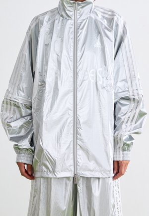 Srebrny, odblaskowy dres Adidas z pełnym suwakiem, elastycznymi mankietami, trzema paskami na rękawach i logo Adidas na piersi.