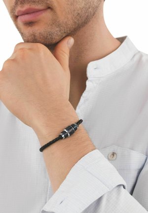 Hombre con barba ligera que lleva una camisa blanca, apoyando la barbilla en una mano que muestra una pulsera trenzada negra con cierre metálico en la muñeca.