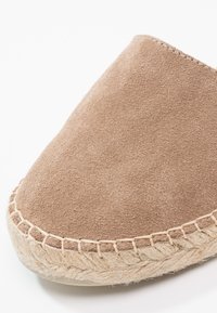 Scarpa slip-on in suede marrone con punta arrotondata, presenta una suola in corda di juta e cuciture visibili lungo il bordo. Superficie texture.